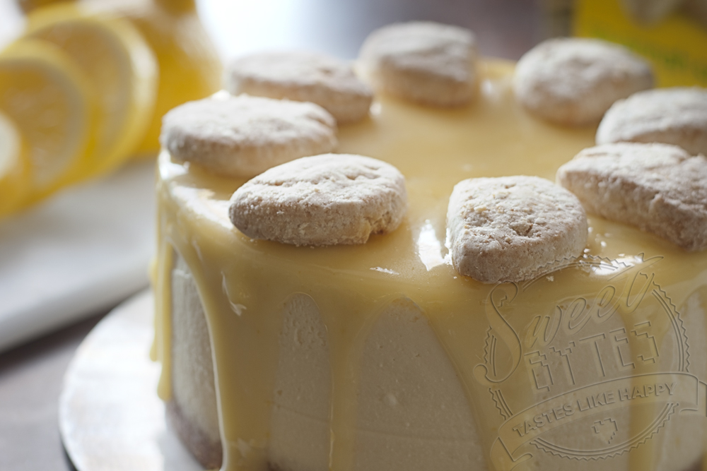 A round lemon cheesecake with yellow lemon curd dripping over the sides. A ring of powder sugar covered Savannah Smiles lemon cookies arranged around the top.