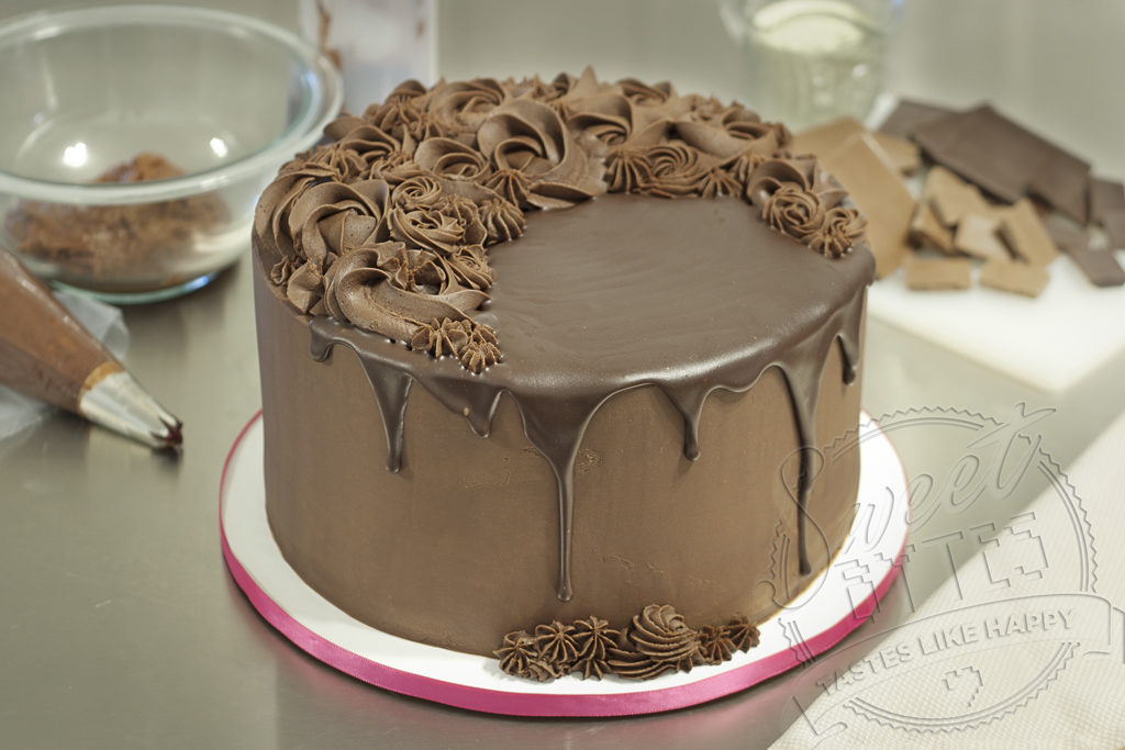 An 8" round cake covered with chocolate ganache frosting. Poured chocolate drips down the sides of the cake and piped chocolate ganache decorations cover the top.