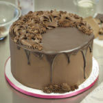 An 8" round cake covered with chocolate ganache frosting. Poured chocolate drips down the sides of the cake and piped chocolate ganache decorations cover the top.