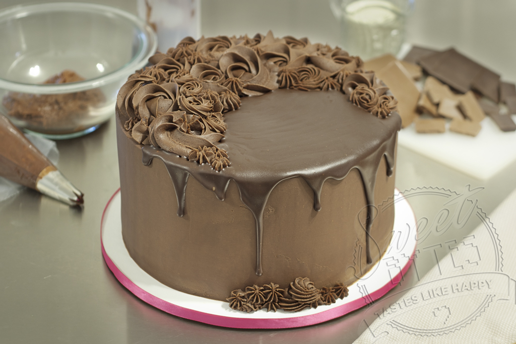 An 8" round cake covered with chocolate ganache frosting. Poured chocolate drips down the sides of the cake and piped chocolate ganache decorations cover the top. 