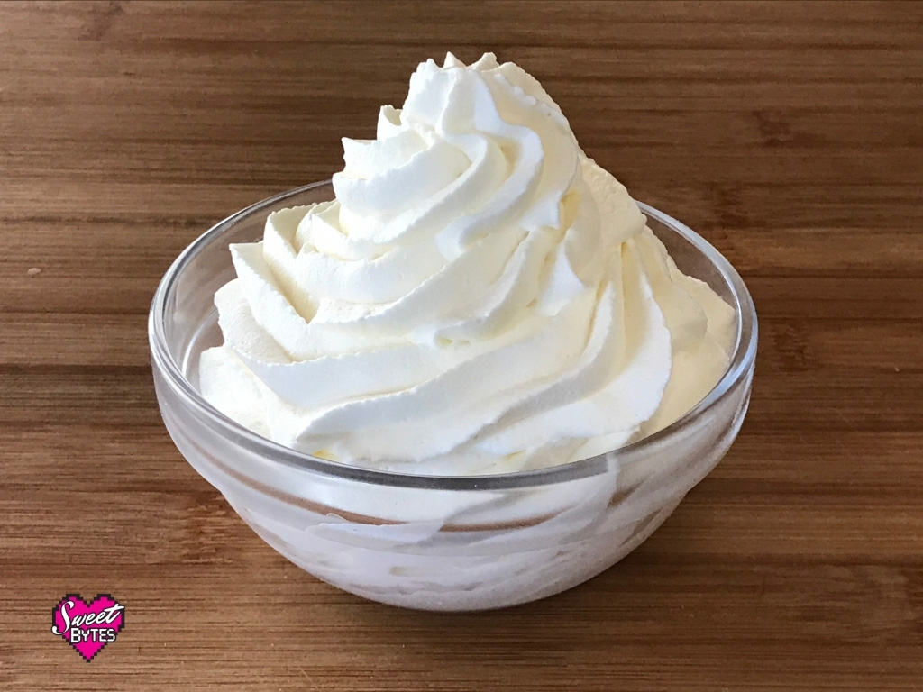 A small glass bowl on a wooden table filled with a heaping swirl of homemade stabilized whipped cream 
