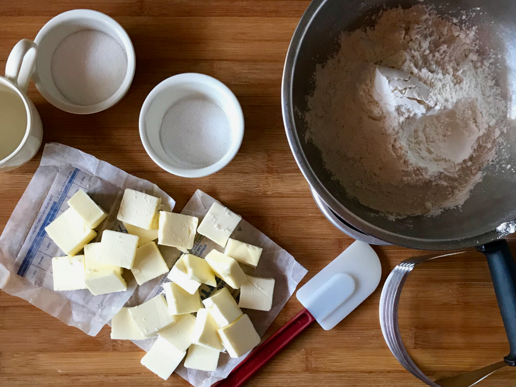 Baking ingredient arranged on a maple cutting board. Cubed butter, 2 white ramekins, a stainless steel mixing bowl with flour