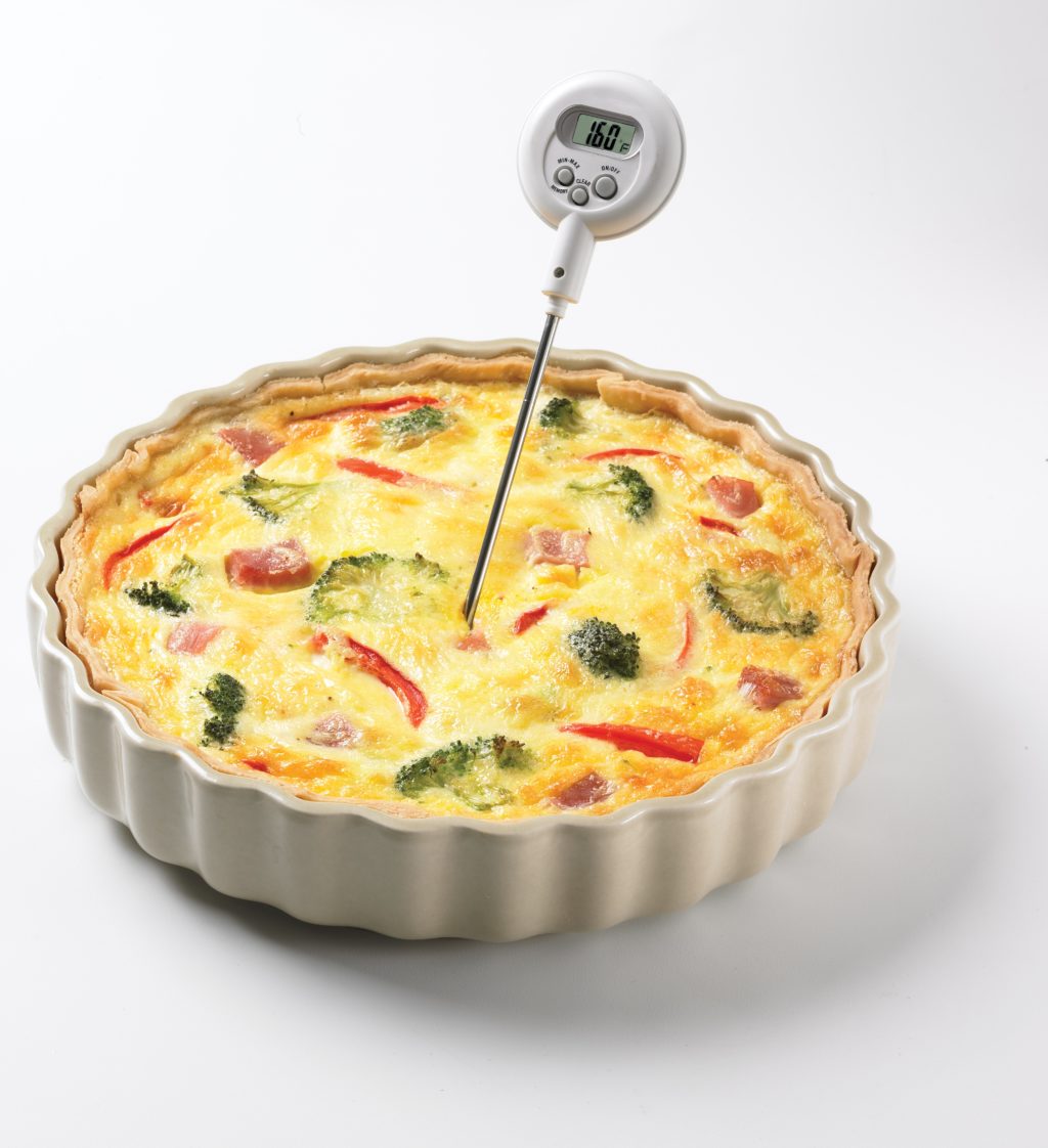 A quiche baked in a tart pan with red and green pieces of bell peppers showing in the yellow egg and cheese mixture. A food safety digital thermometer sticks up from the center of the quiche. 