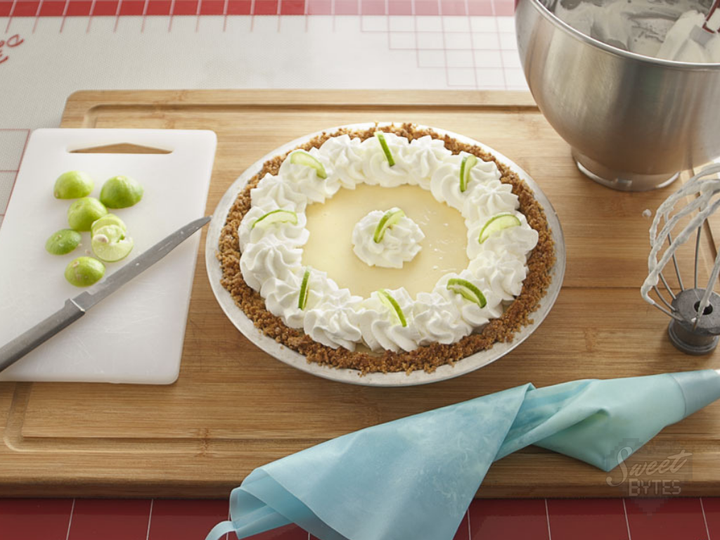 A key lime pie garnished with whipped cream and sliced key limes sitting on a wooden cutting board