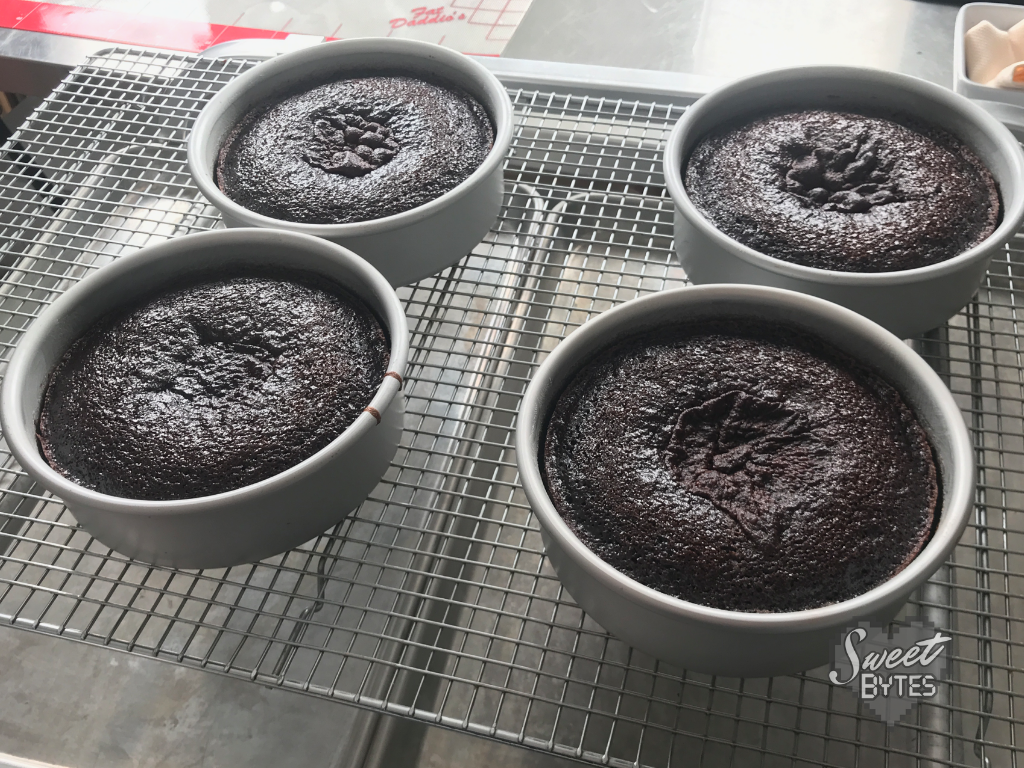 Four 6" chocolate cake layers in Fat Daddio's pans cooling on a cooling rack