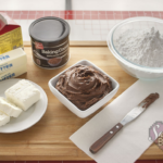 A small, white, square bowl of chocolate cake frosting surrounded by the recipe's ingredients