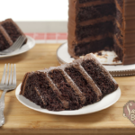 Four layer chocolate cake slice on white dessert plate with the sliced chocolate cake behind it