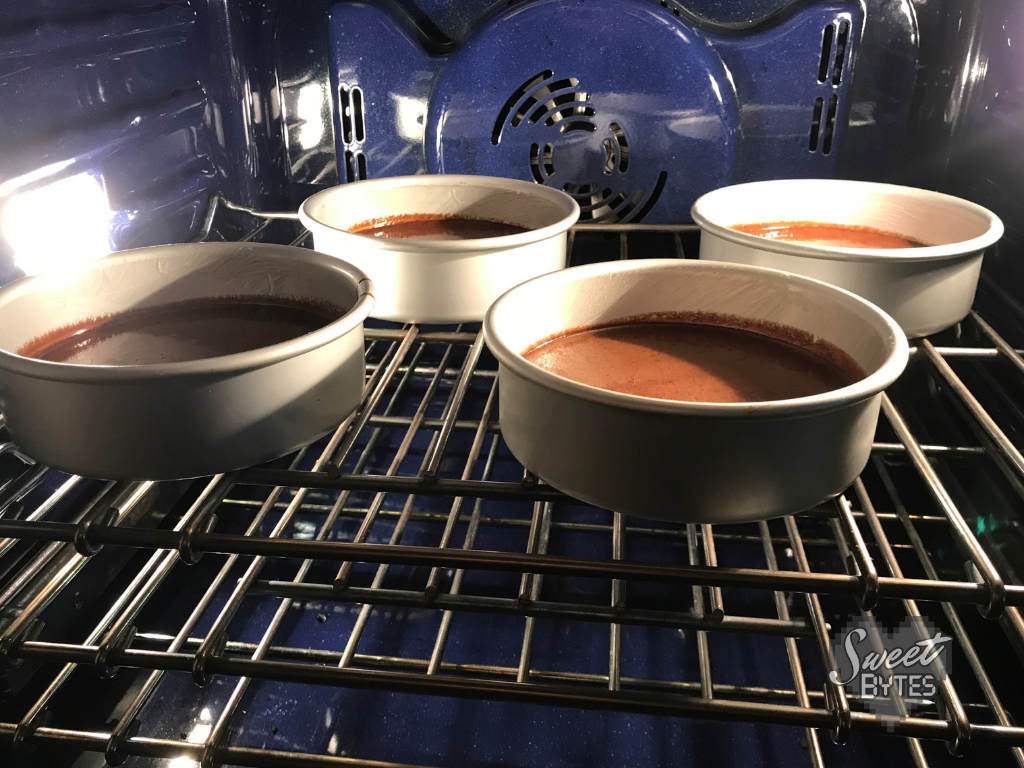 Make sure there is 1" of space between your cake pans and the oven wall for the best cake baking results. #sweetbytesokc #howtobake #cake #baking #recipe #chocolatecake Four pans filled with chocolate cake batter inside of an oven with blue walls