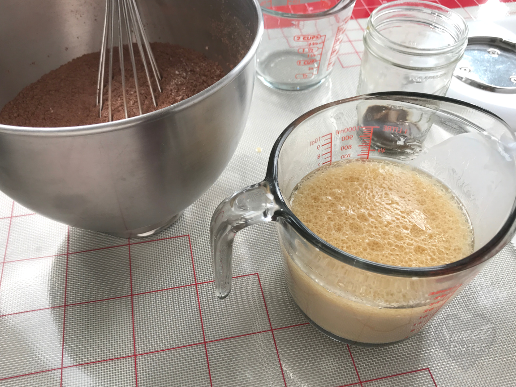 A stainless steel mixer bowl with well mixed dry ingredients for a chocolate cake and a glass measuring cup with all wet ingredients for the chocolate cake mixed together