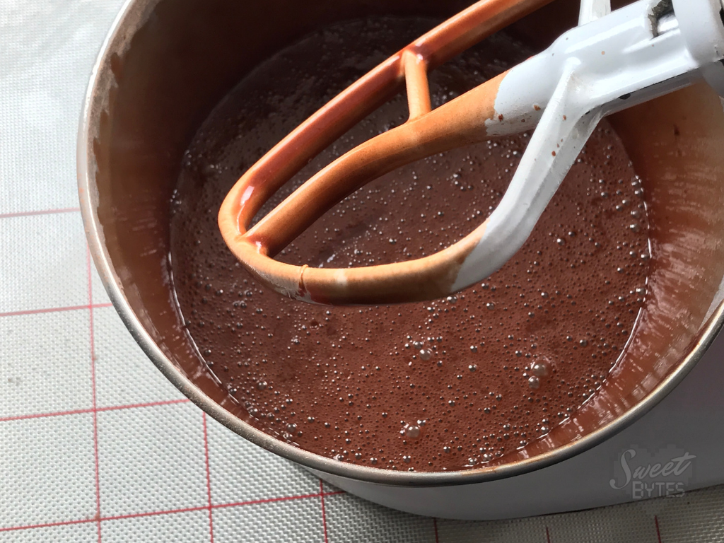 Close up of finished chocolate cake batter in a stainless steel mixer bowl with stand mixer paddle attachment covered in thin cake batter showing