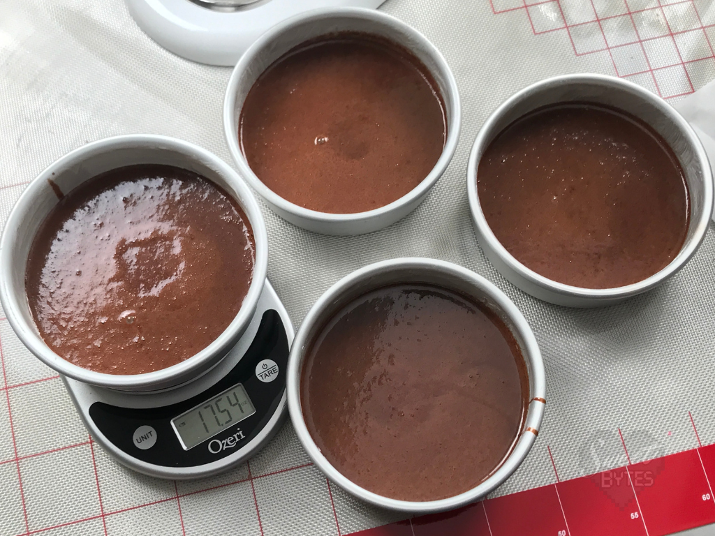 Overhead shot of 4 six inch cake pans filled with chocolate cake batter. The far left pan is sitting on a kitchen scale showing 17.54 ounces