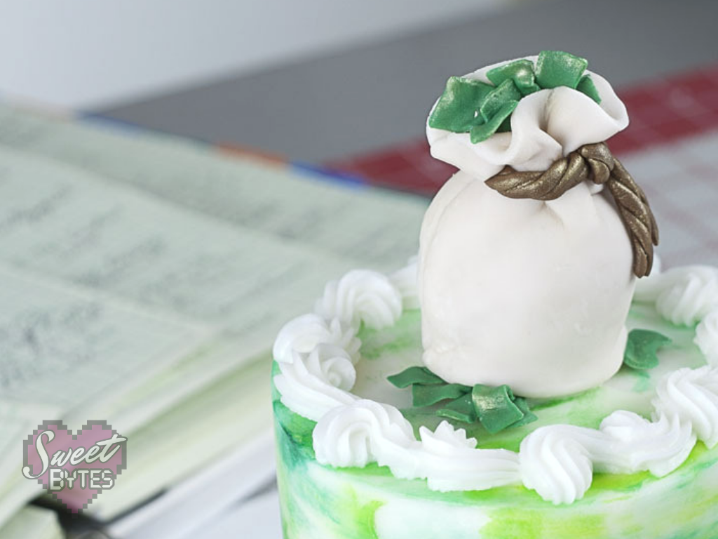 top of a small green and white cake with a fondant money bag cake topper with green fondant money coming out of the top and bottom for the costs of bakery items