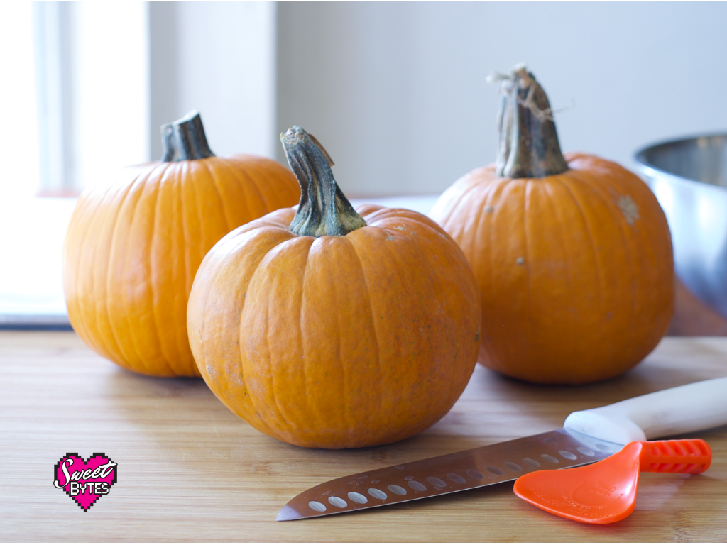 Wondering how to cook pumpkin for pie? This article will show you how! 3 pie pumpkins sitting on a wooden table with a large knife and pumpkin scraping tool.