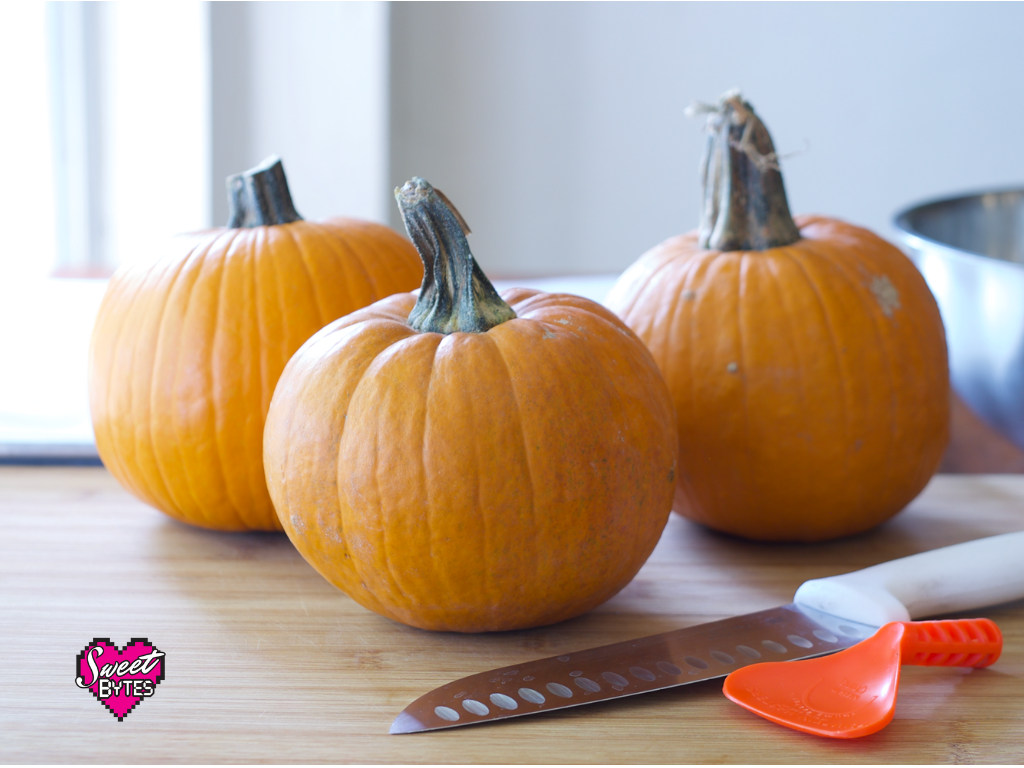 3 pie pumpkins sitting on a wooden table with a large knife and pumpkin scraping tool.