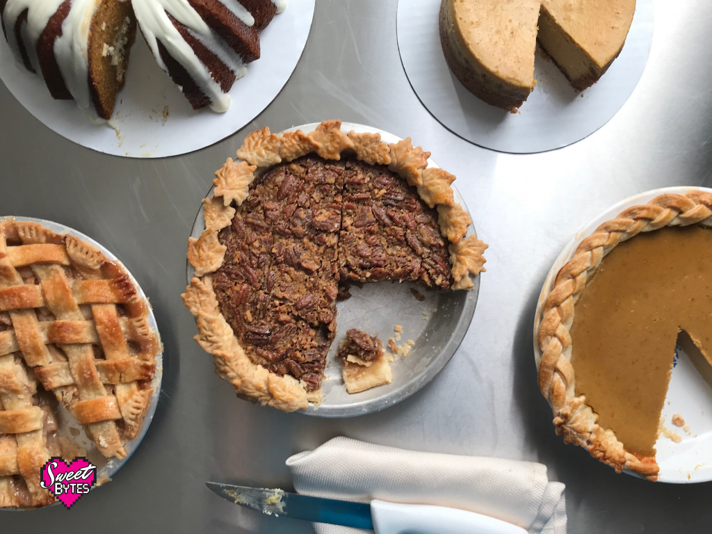 Overhead shot of Thanksgiving desserts on a stainless steel tabletop. Caramel apple pie, pumpkin bundt cake, bourbon pecan pie, pumpkin cheesecake, pumpkin pie