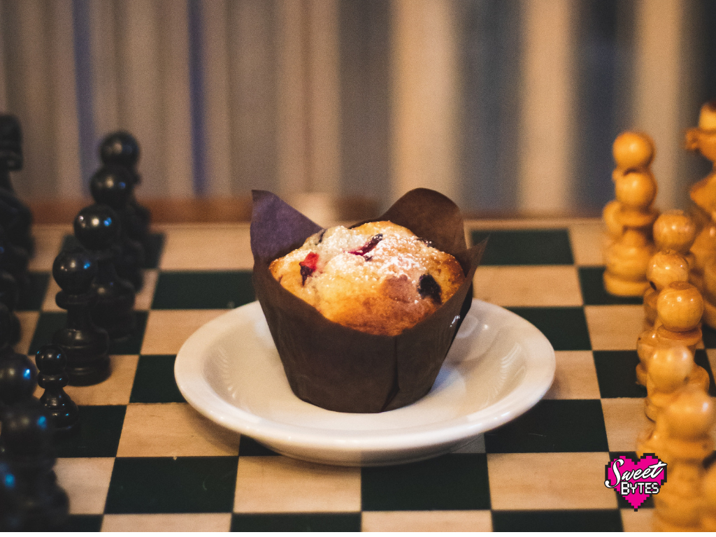 a muffin in a brown tulip style wrapper on a white saucer on the middle of a chess board