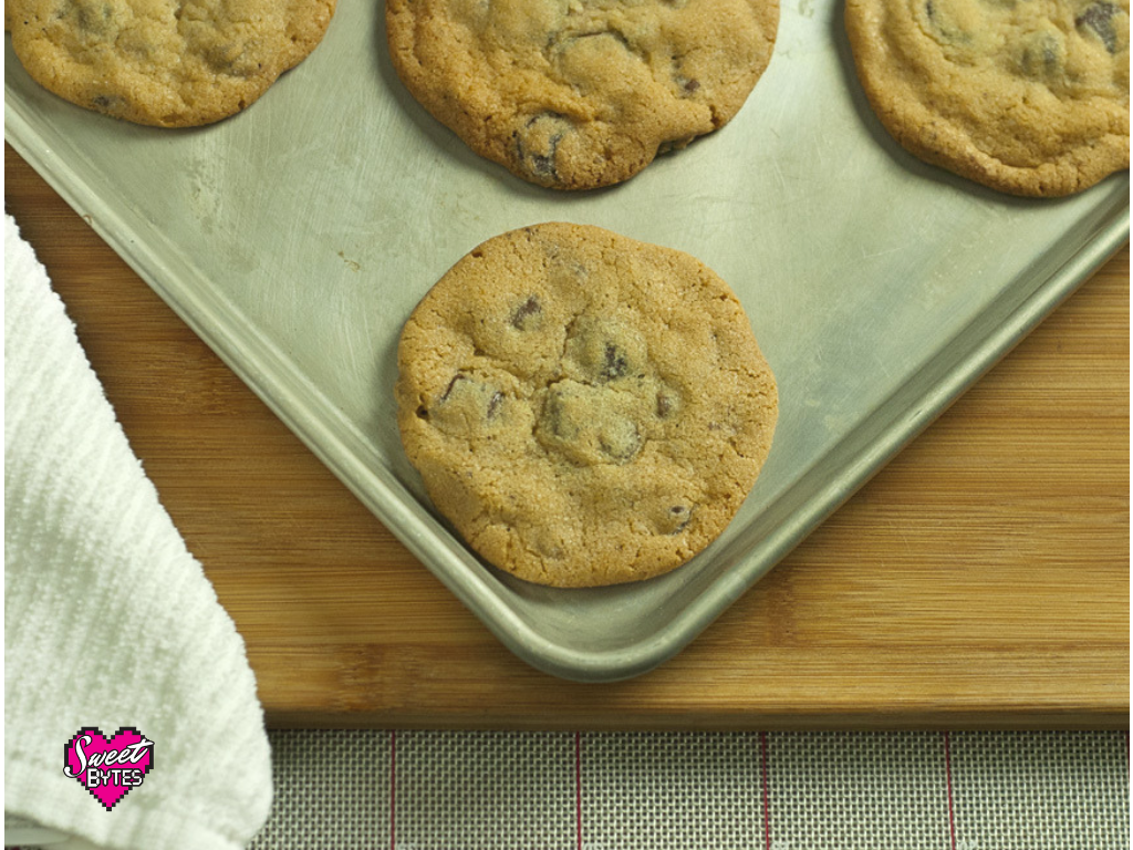 Close up view of baked chocolate chip cookies on a baking sheet sitting on a cutting board with a white towel near the bottom left edge