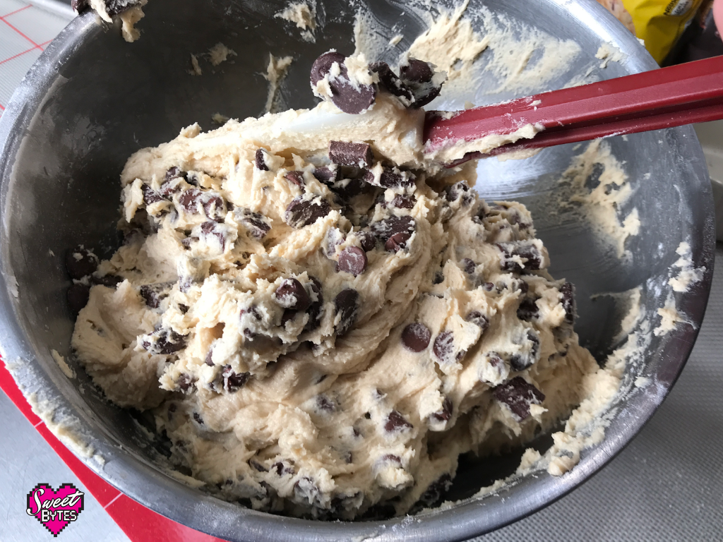 A stainless steel bowl of chocolate chip cookie dough, chocolate chips being blended in by hand