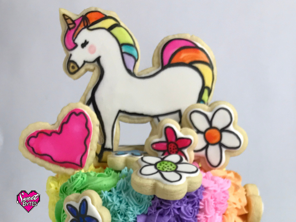 A handpainted unicorn sugar cookie, heart cookie, and flower cookie on top of a cake