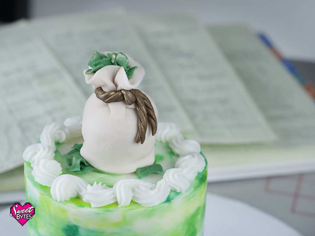 small cake with a money bag cake topper with green and white buttercream frosting in the foreground with an open ledger in the background to show home bakers how to pay themselves