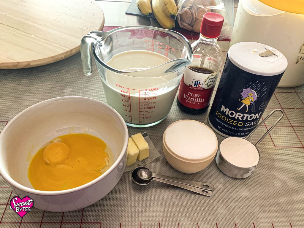 Ingredients for making vanilla pudding pie filling laid out on a grid lined silicone mat. A white bowl with egg yolks, a measuring cup with milk and cream, a measuring cup with sugar, vanilla extract and a container of Morton's salt. 