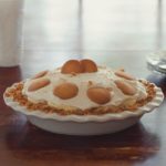 Homemade Banana Pudding Pie in a white ceramic LeTauci pie plate sitting on a wooden table with a window and white dishes in background.