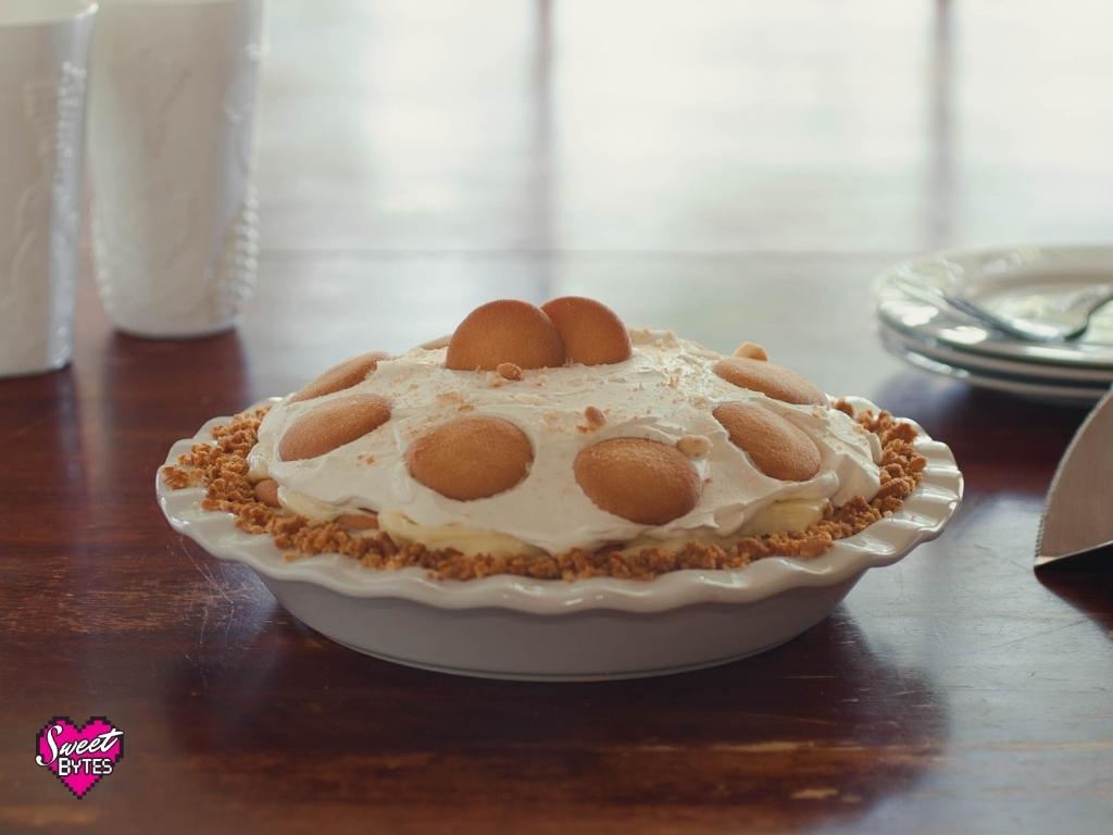 Homemade Banana Pudding Pie in a white ceramic LeTauci pie plate sitting on a wooden table with a window and white dishes in background.