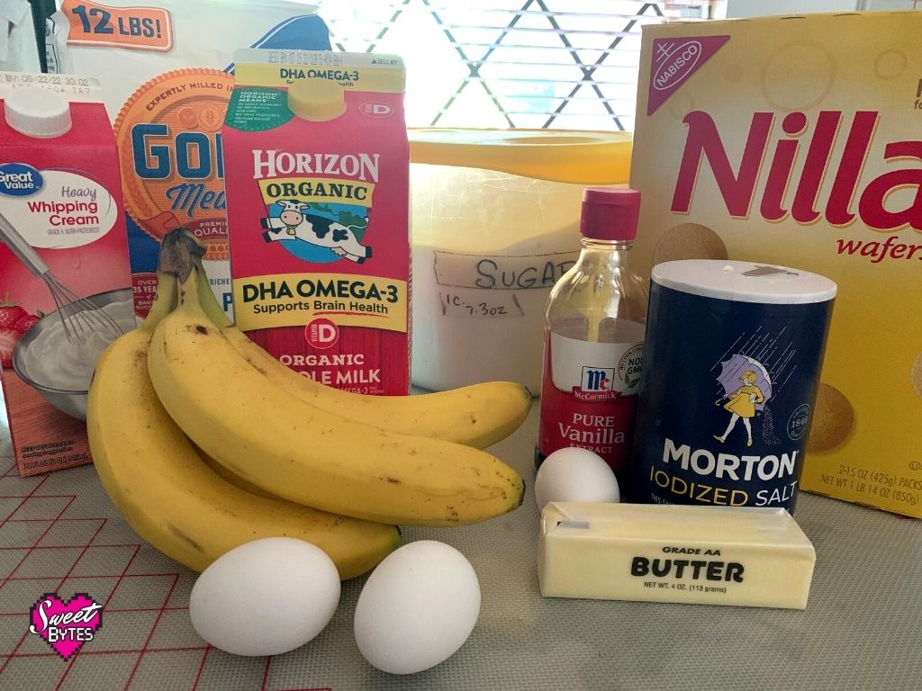 Ingredients for homemade banana pudding pie on a table with a window in the background. From left to right the ingredients shown are heavy cream, bananas, all purpose flour, eggs, milk, sugar, butter, vanilla, vanilla wafers, and salt.
