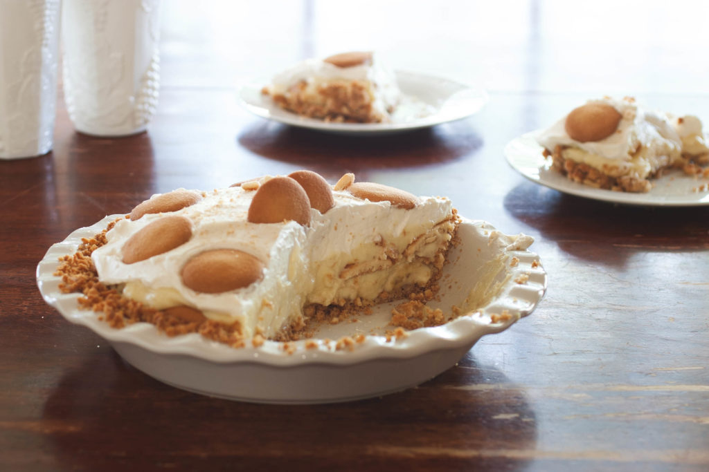 Banana pudding pie in a white pie plate with wavy edges. There are two slices of pie missing from the whole pie, The slices are on white saucers behind the pie. Sunlight is streaming through the window onto the scene.