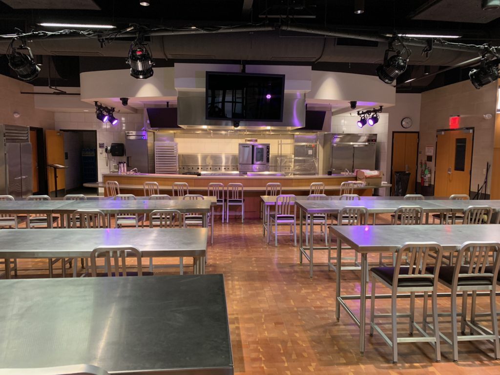 Stainless steel tables and aluminum chairs set up in a class room style. Kitchen area and large display screens are in the background, studio lights are hanging from the ceiling in this state of the art demonstration kitchen classroom at Francis Tuttle in Oklahoma City, Rockwell Campus.