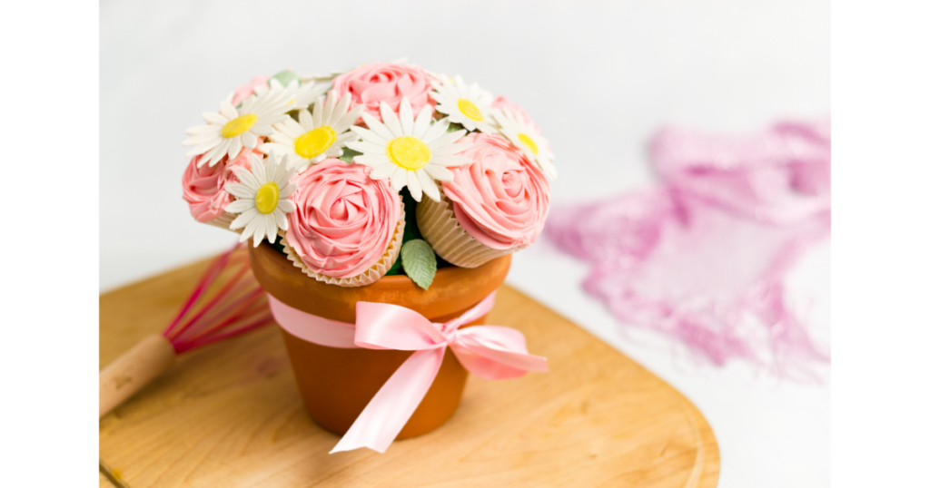 A cupcake bouquet set in a terra cotta pot with pink rosette cupcakes and white daisies made of paper on wooden cutting board